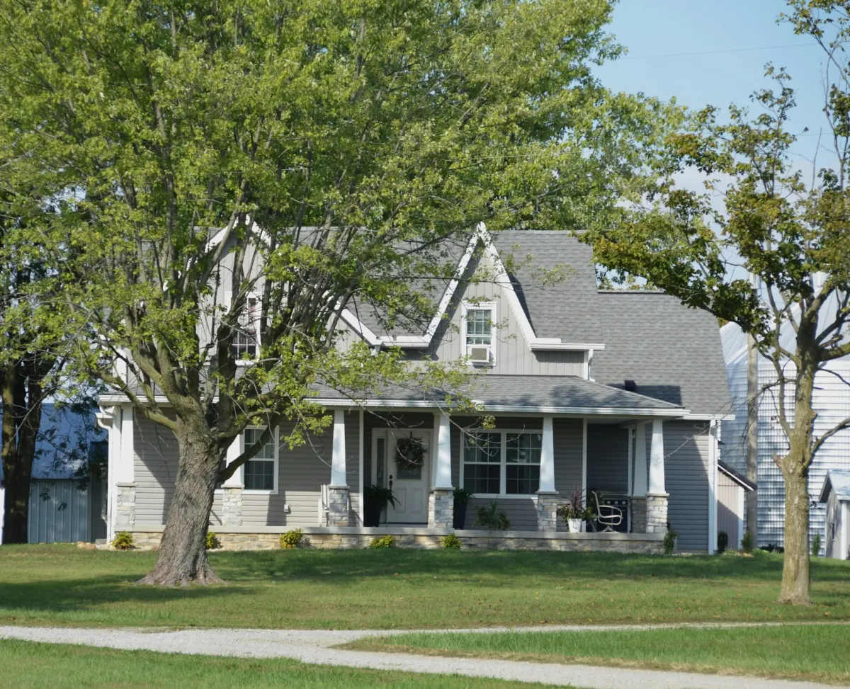 A gray farmhouse with a covered porch set back on a lawn behind mature trees