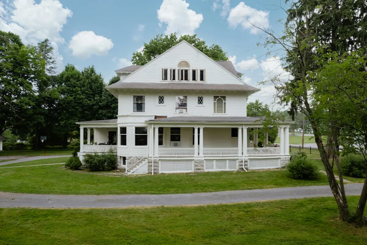 A large older white house with a wraparound porch, mature trees and an open lawn