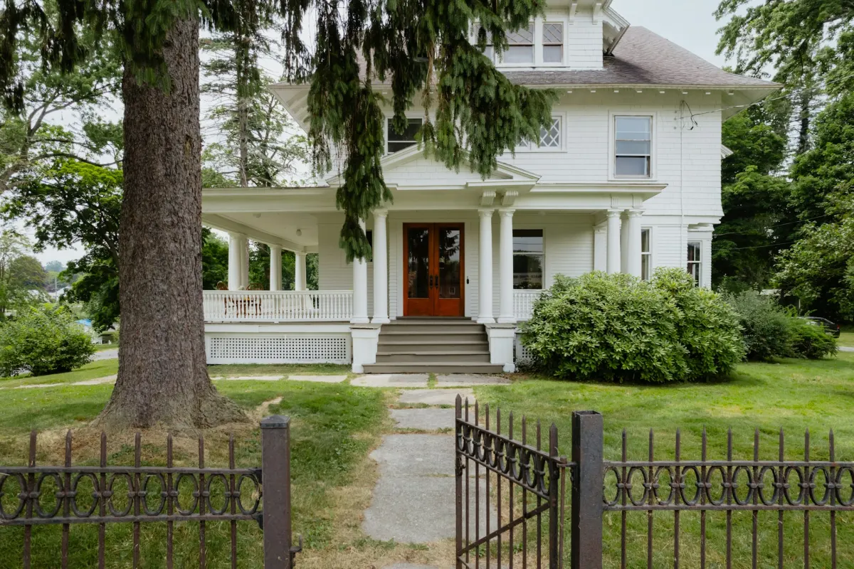 An older white two-story house with a columned front porch behind an iron fence